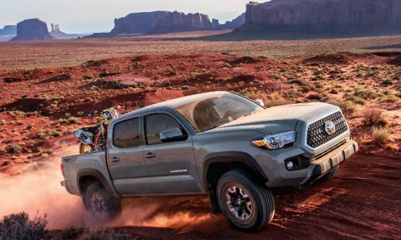Cement Gray 2018 Toyota Tacoma Climbing Hill in Desert with Dirt Bike in Bed and Rock Formations in Background
